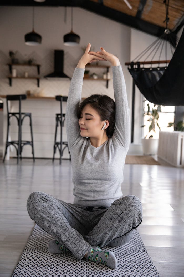 about-01 Cheerful young ethnic female in earbuds sitting on yoga mat with crossed legs and closed eyes while listening to music and raising hands in apartment