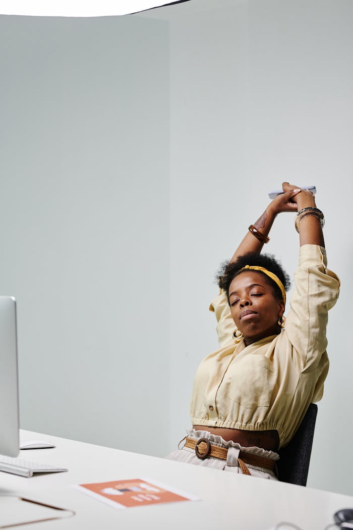 gallery-6 African American woman stretching with eyes closed at her office desk, feeling relaxed.
